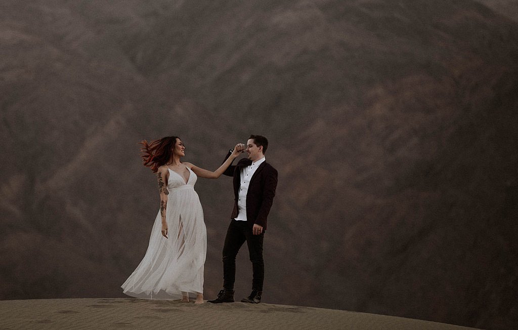 Adventure elopement in Oregon wedding couple dances on top of the mountain in the wind. Tattooed bride and lace dress for hiking elopement