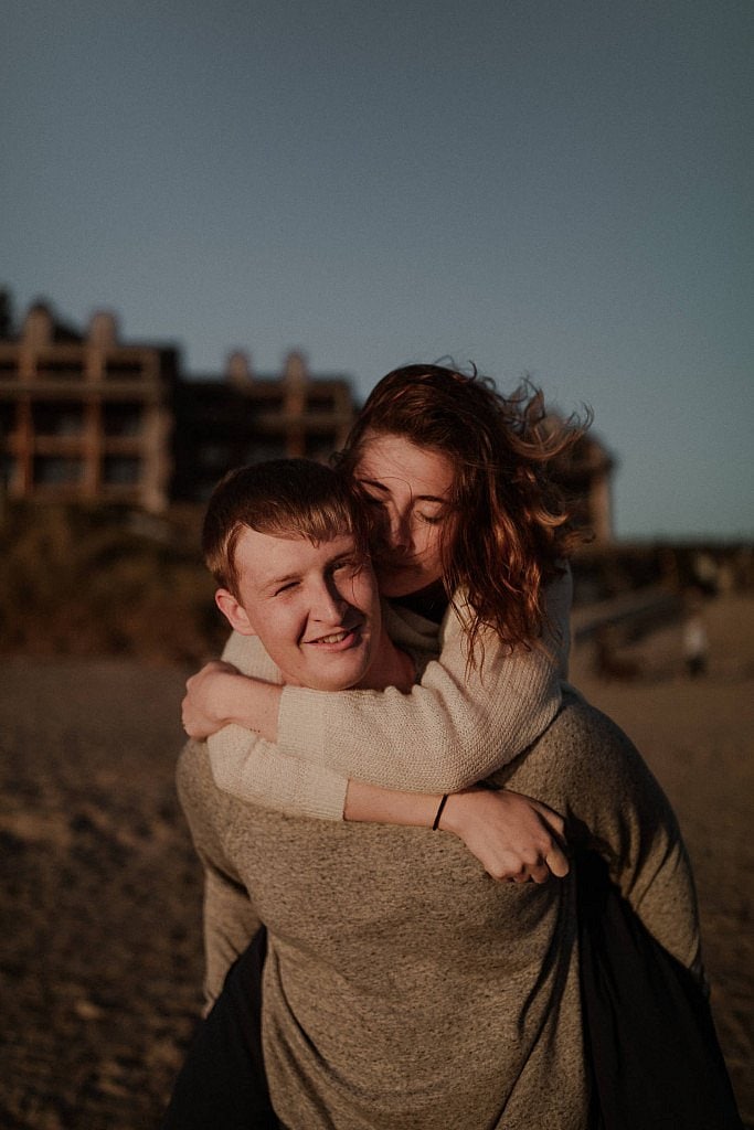 Bride clutches her groom from behind as he carries her through the sand. Romantic Oregon coast beach elopement at sunset and the light makes their skin glow