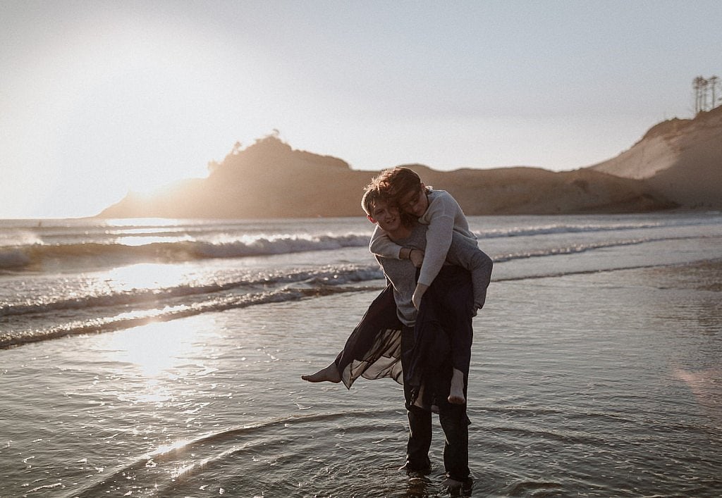 Groom carries his bride into the cool water after their elopement ceremony. Cape Kiwanda on the moody Oregon Coast is a perfect spot to get married on the beach