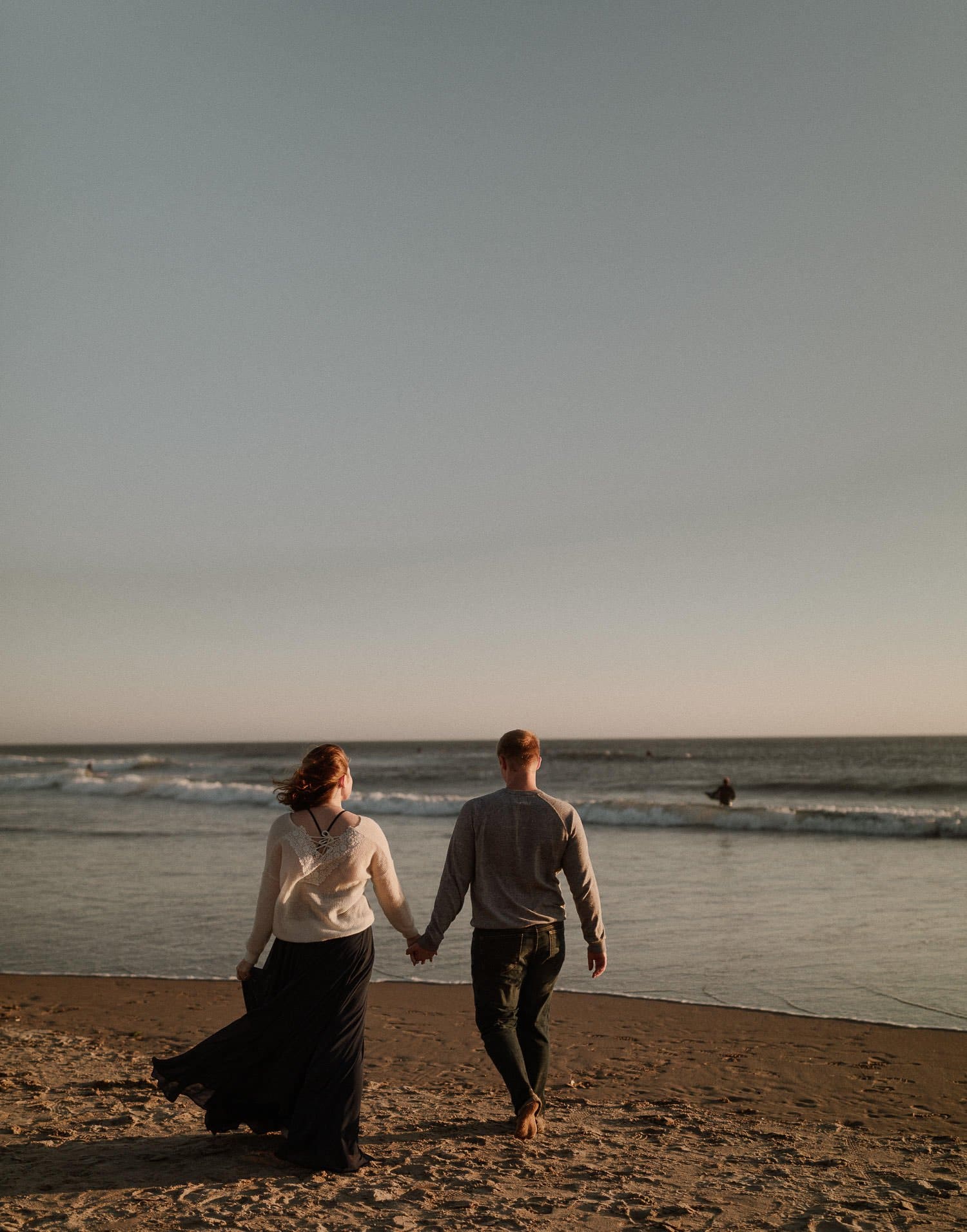 Groom holds brides hand as they walk along the beach at sunset after their elopement ceremony. The waves slowly roll over the sand