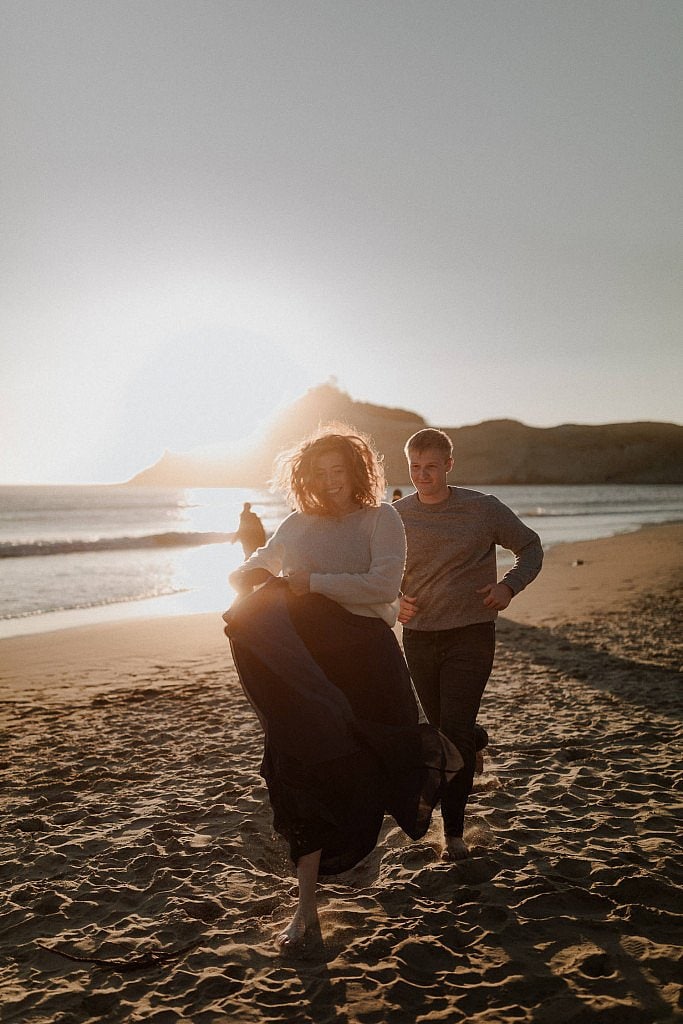 Bride and groom run across the beach in a game of tag. Bride holds her flowy skirts up and her red hair glows in the evening light
