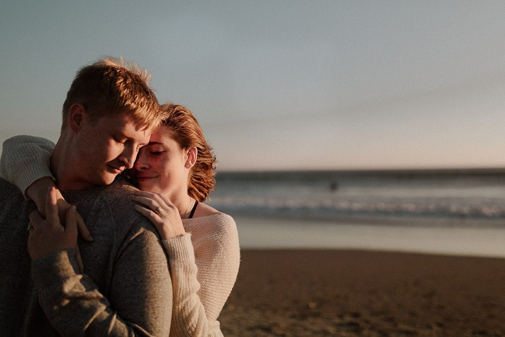 Bride cuddles next to her groom and the sunset lights half of their faces in the light. Their cozy sweaters keep them warm against the ocean breeze