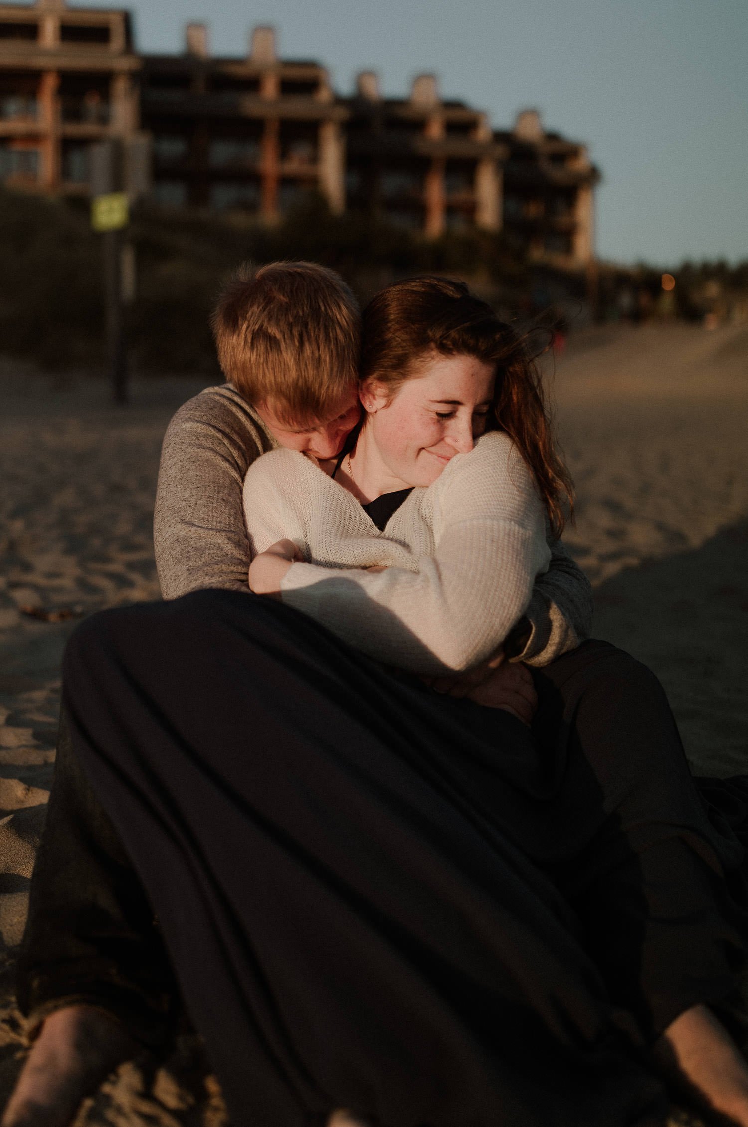 Bride snuggles closely into groom so he can keep her warm while they sit on the ground in the sand after their beach elopement at Pacific City Beach