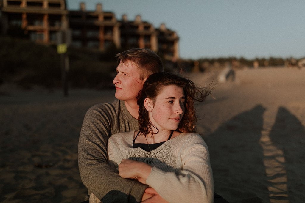 Bride and groom sit on the warm sand and cuddle after their intimate elopement ceremony on the beach. The setting sun gently lights their faces as they look in opposite directions