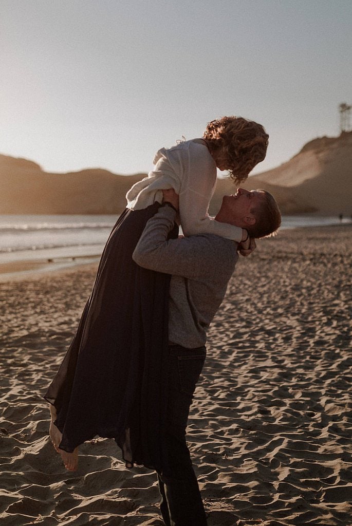 A groom picks his bride up at the waist under her white sweater and carries her over the sand for their Cape Kawanda Beach elopement on the Oregon Coast