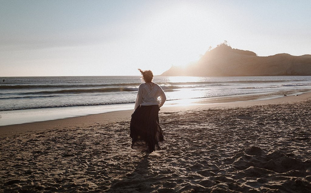 A bride runs down the beach at sunset on the Oregon coast with her navy blue flowy wedding dress and a white sweater to keep warm. The setting sun makes her hair glow before she touches the cool water.