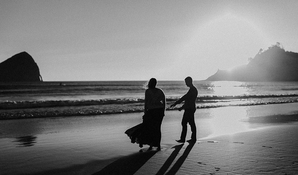 A bride and groom walk the edge of beach along the water at Cape Kiwanda during their sunset coastal elopement in Oregon. Groom reaches his hand out for the bride as she carries her dress so it doesn't get wet