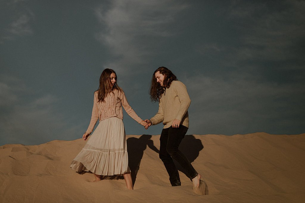Cute sand dune couples session at sunset. Couple climbs up sand dune for adventure session. They wear cozy sweaters and a pink skirt