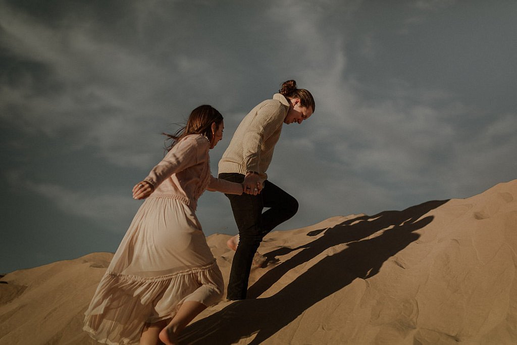 Sand dunes couple session, couple climbs up the tall sand dune with her hair blowing in the wind