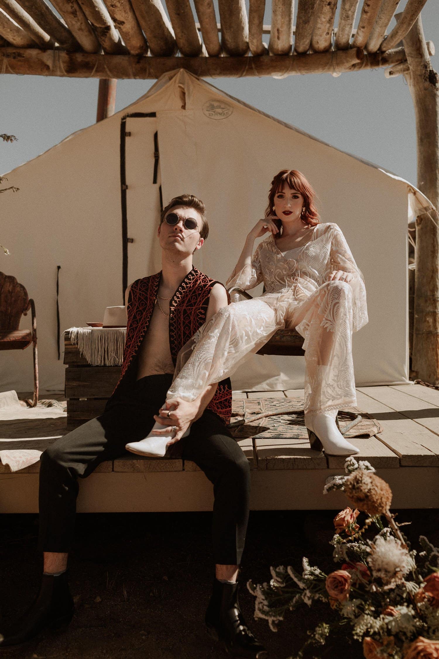 Edgy elopement couple sit outside their desert yurt before their elopement ceremony. Bride wears a bridal jumpsuit and the groom looks cool in his sunglasses