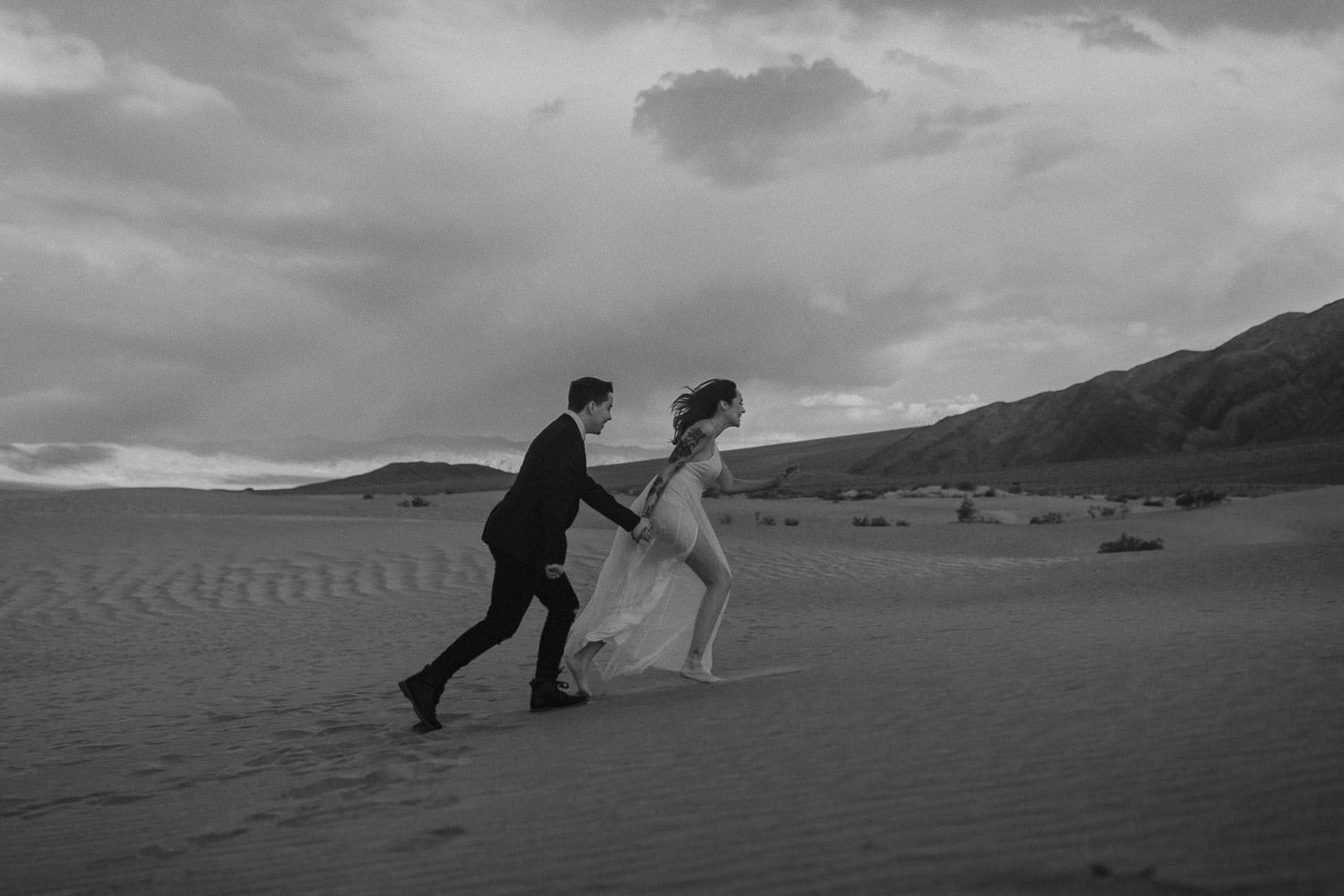 Amazing Oregon desert elopement in the sand dunes. The wedding couple runs up the sand dune in this black and white image. Her elopement dress flows out behind her.