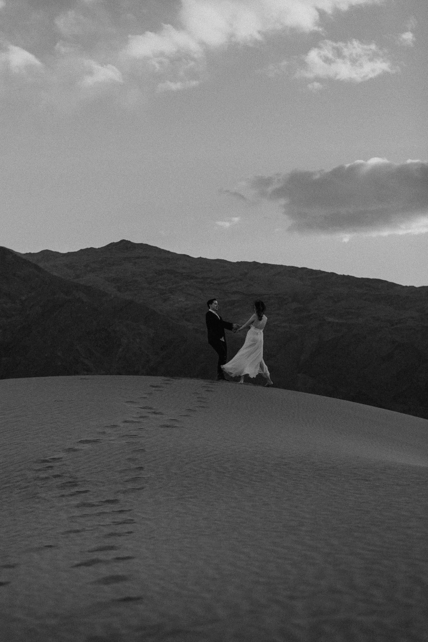 Black and white image of bride and groom dancing at the top of a dune. Their foot prints show in the sand while the groom spins his new bride around