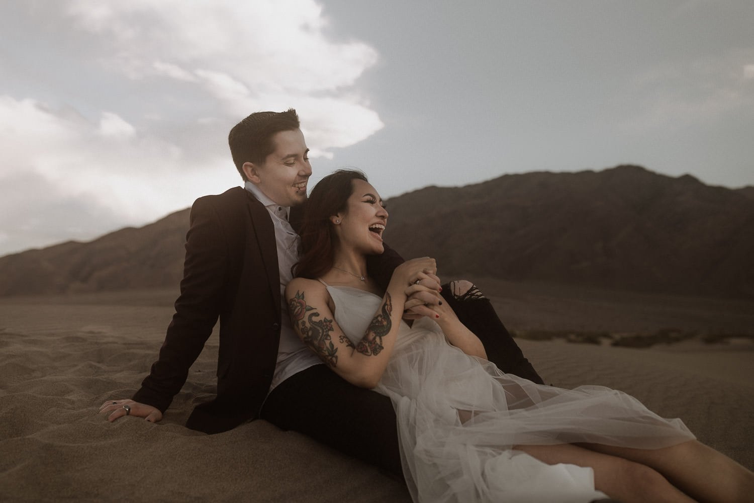 Bride laughs while she leans against her husband. Her dress folds around her feet, perfect for their adventure Oregon elopement in the desert. The. mountains and clouds frame the background