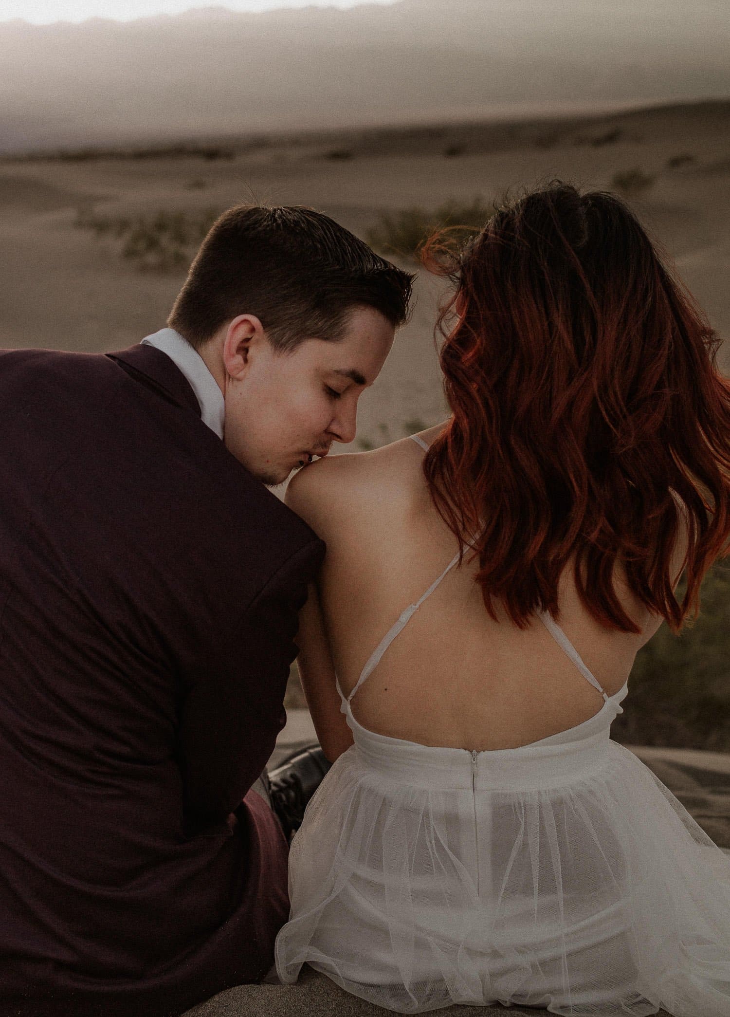 Elopement couple snuggles in the sand at sunset after their intimate elopement ceremony. Groom kisses her shoulder gently