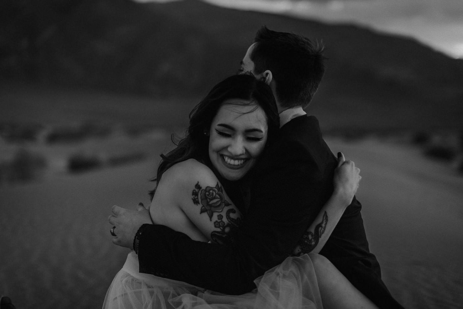 Black and white image of bride and groom laughing and snuggling on the ground. Tattooed bride snuggles closely to him to keep warm from the wind