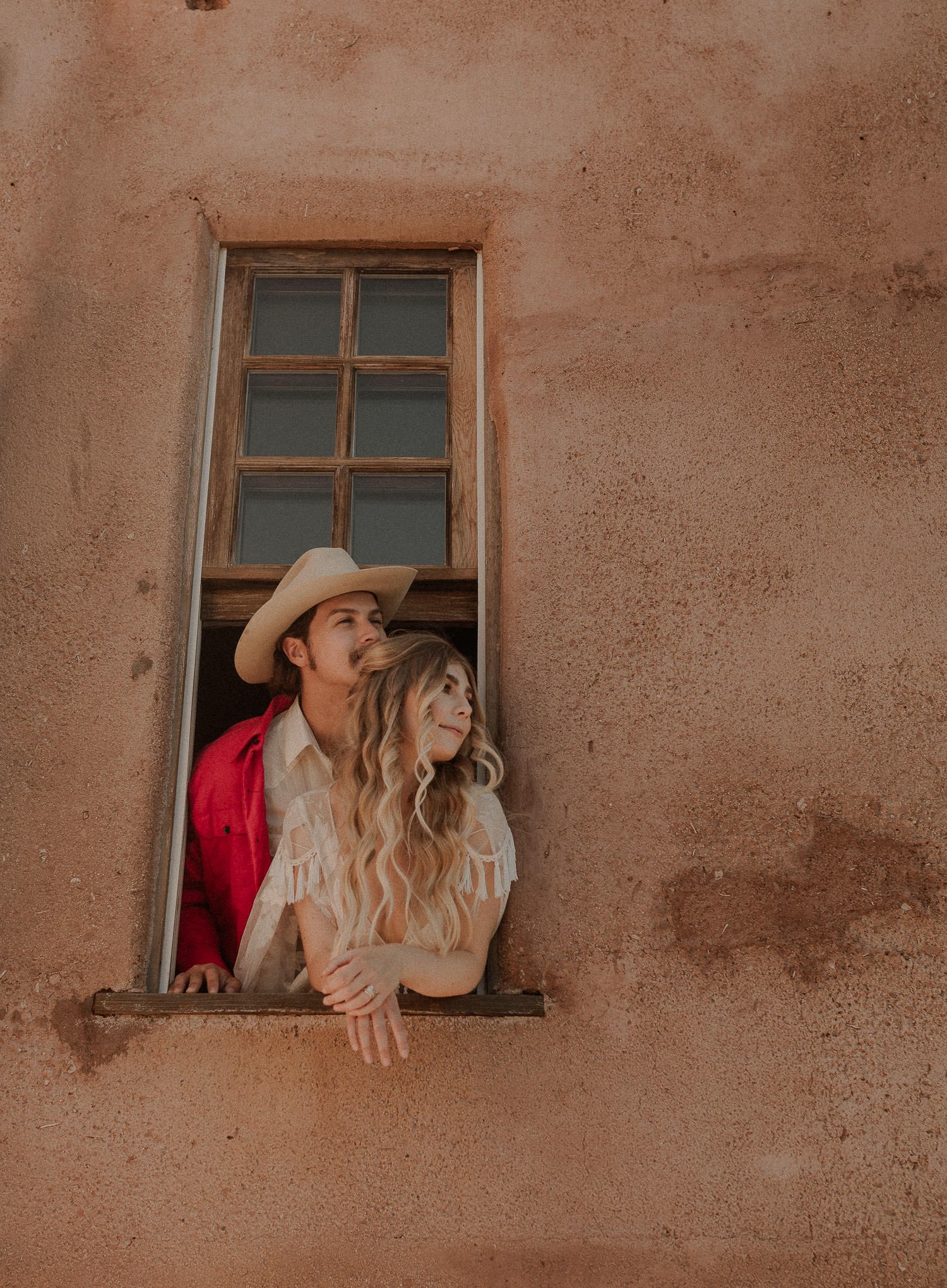 Bride and groom hang out of window on vintage adobe in the desert. Bride's long blond hair looks like rapunzel and the groom wears a bride red jacket and cowboy hat