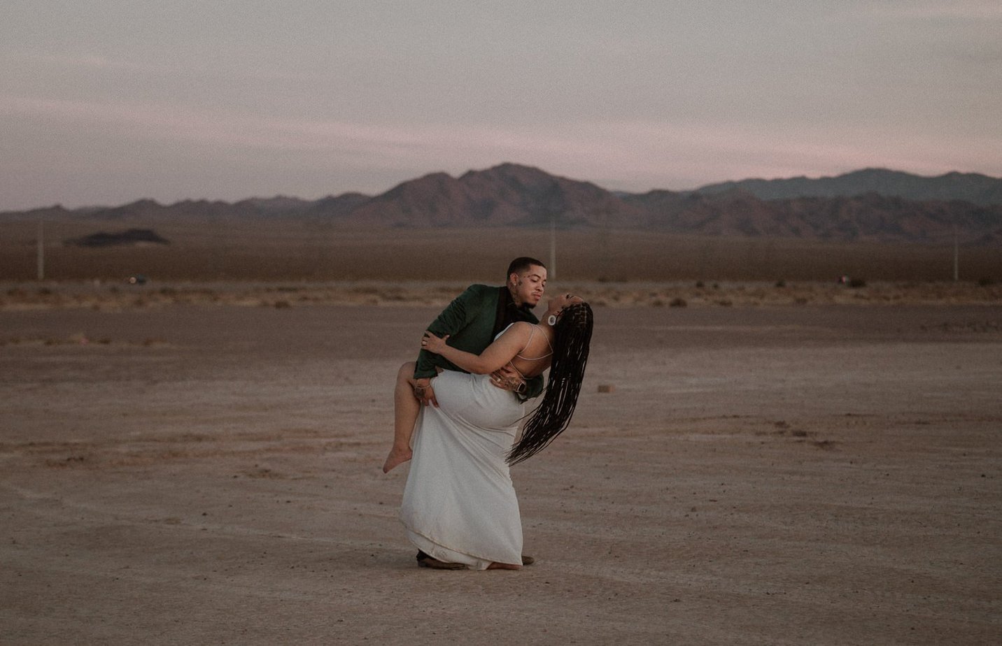 Fun desert elopement by Oregon elopement photographer Black Salt Photography. The mountains look purple behind the couple as the sun sets. Groom dips his bride back and her long hair blows in the wind