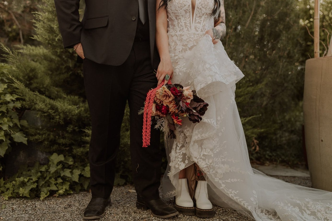 Bride and groom stand next to each other. Edgy bride shows off her Doc Martin boots for her wedding shoes and holds her bouquet by her side.