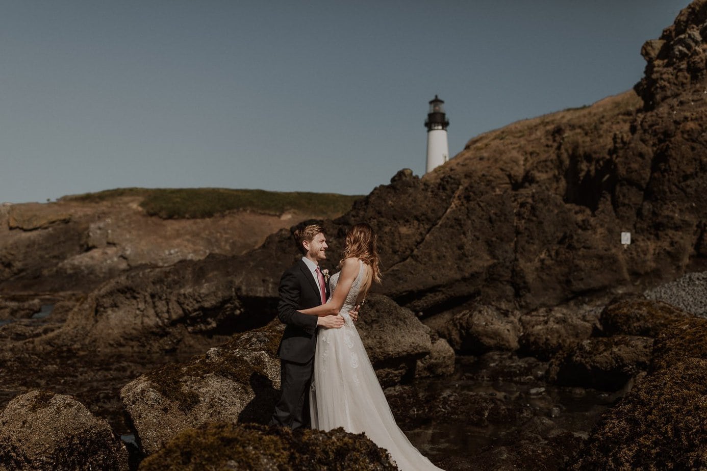 Romantic lighthouse elopement on Cobble Beach. The Oregon coast is full of amazing places to elope. Wedding couple caresses on the black stone beach in front of Yaquina Head Lighthouse