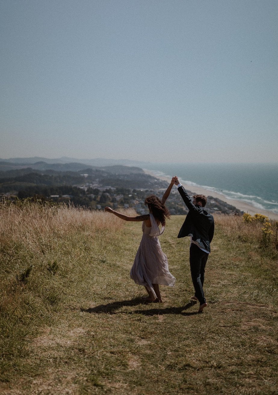 Amazing Oregon coast elopement at God's Thumb. Elopement couple dances on the grassy hillside, her lavender elopement dress blowing in the wind. The blue waters show the shoreline in the distance