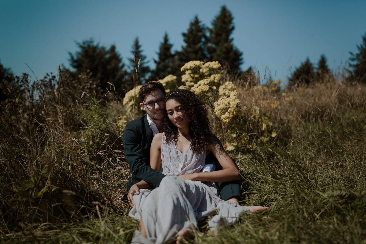 Bride and groom sit on the ground surrounded by yellow wildflowers and grass. The sun is bright on their faces. They both are barefoot for their adventure elopement