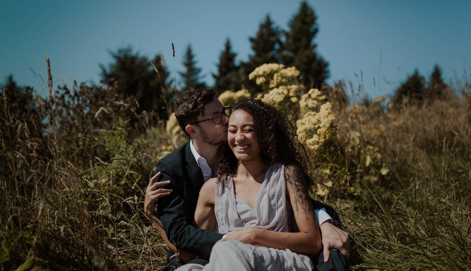 Adorable eloping couple snuggle on the ground among the wildflowers before their God's thumb elopement on the Oregon coast. The bride wears a lavender wedding dress and the groom has a dark blue suit on. The groom kisses the side of her cheek and she laughs happily