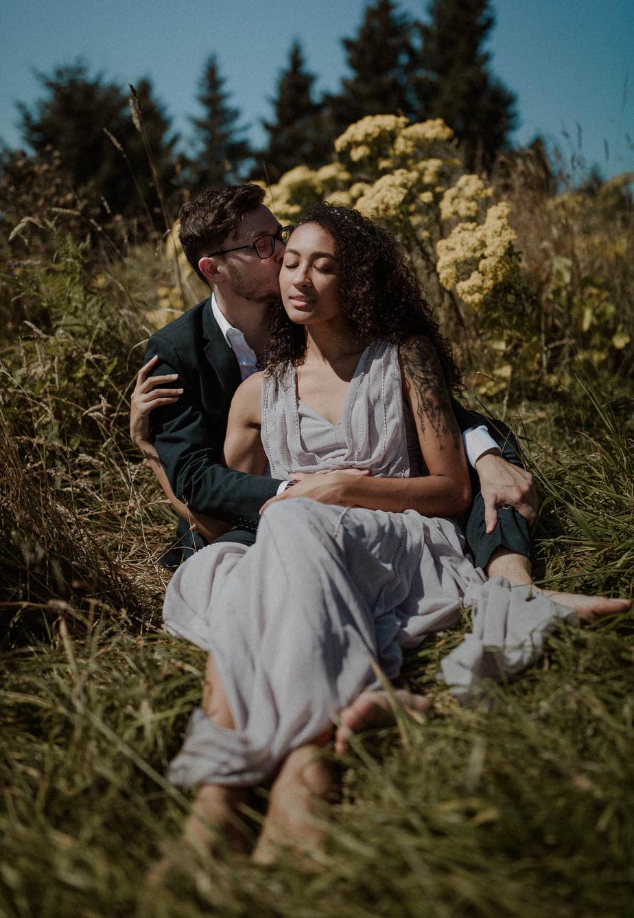 God's Thumb Elopement, Bride and groom sit on the grass surrounded by yellow wildflowers at their God's thumb elopement on the Oregon coast. Bride wears a lavender wedding dress and bare feet. Groom kisses the side of her face