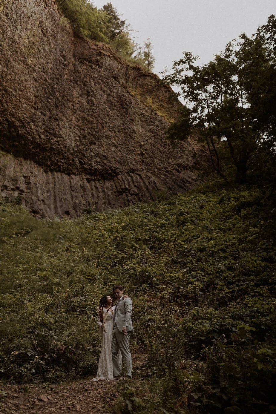 Bride and groom stand at the edge of a forest for their Latourell Falls elopement. The large canyon above them makes them look small