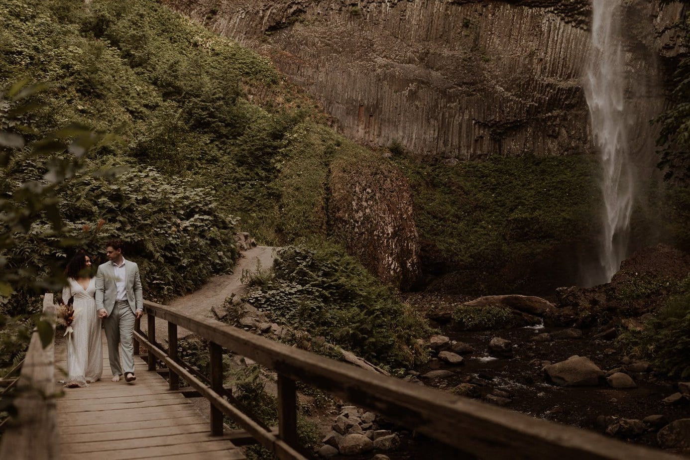 Wedding couple walks across wooden bridge at Latourell Falls for their Oregon Waterfall elopement. The waterfall is in the background
