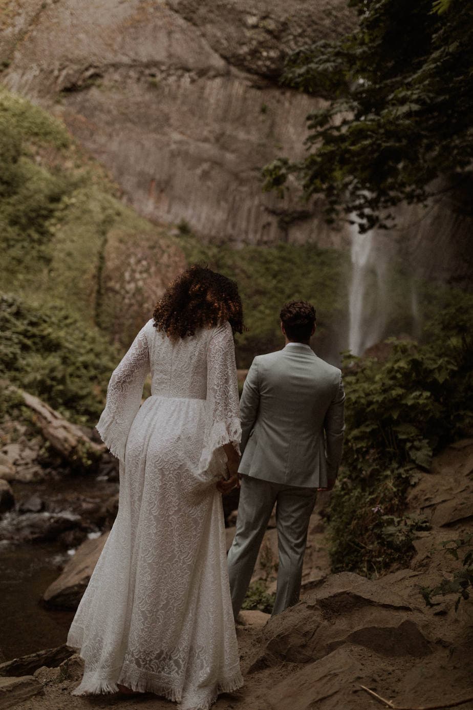 Latourell Falls Elopement Bride and groom walk across the rocks for their waterfall elopement in Columbia River Gorge. The Gorge is full of so many waterfalls that you can visit but Latourell falls is one of the easiest to get to