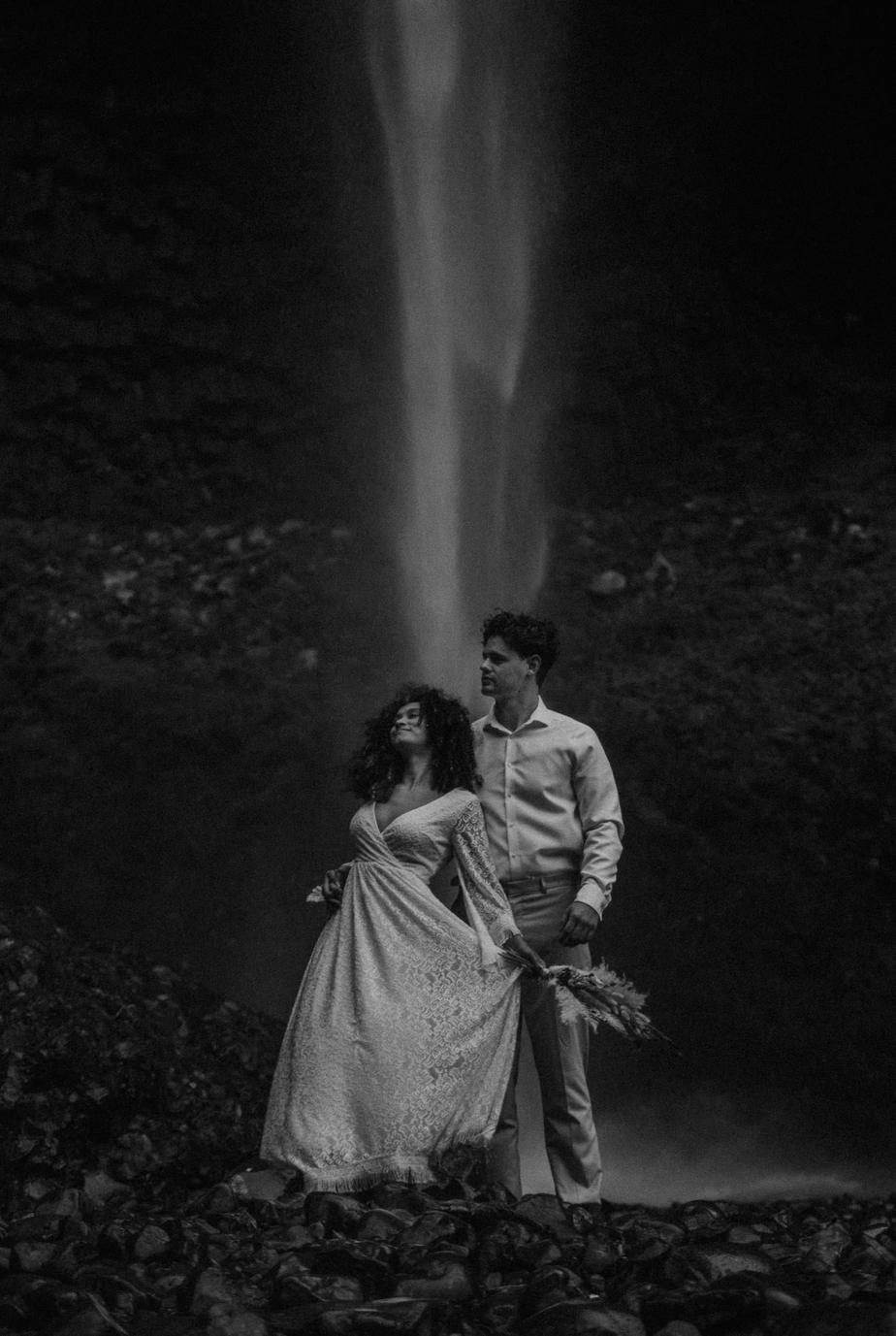 Black and white image of bride and groom standing together in front of Latourell Falls in the Columbia River Gorge in Oregon. The breeze blows the brides boho dress and her natural hair