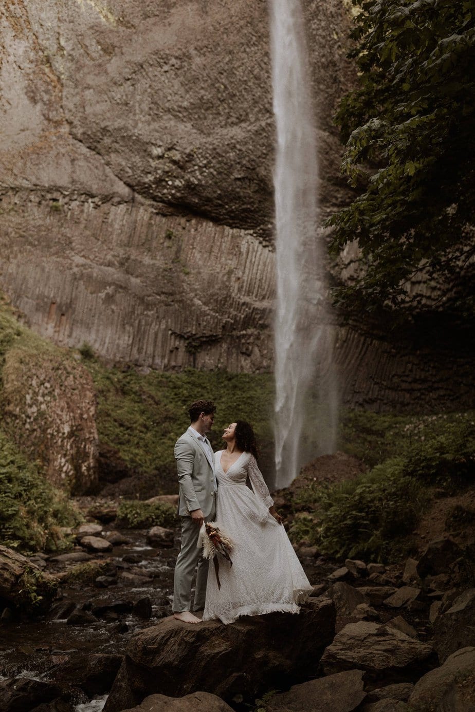 Wedding couple look up at each other on top of a large rock in front of a waterfall. Latourell falls is the perfect place for your elopement