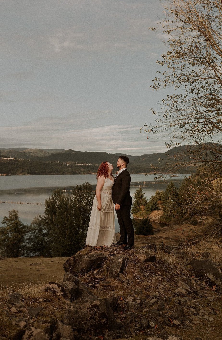 Wedding couple stand on cliff at Government Cove elopement. The sunset casts a pretty glow on their skin. Bride looks up at her groom while he holds her. The river and mountains are in the background.