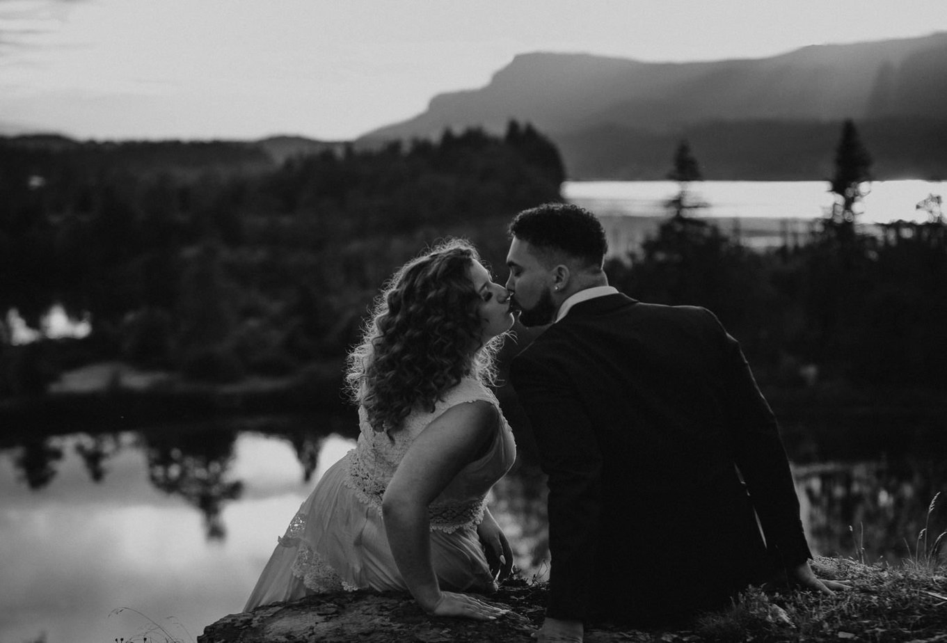 Government Cove Oregon adventure elopement in the Columbia River Gorge. Black and white image of couple sitting on the edge of the cliff at sunset. The mountains and trees reflect in the rivers surface. Bride leans in to kiss the groom gently