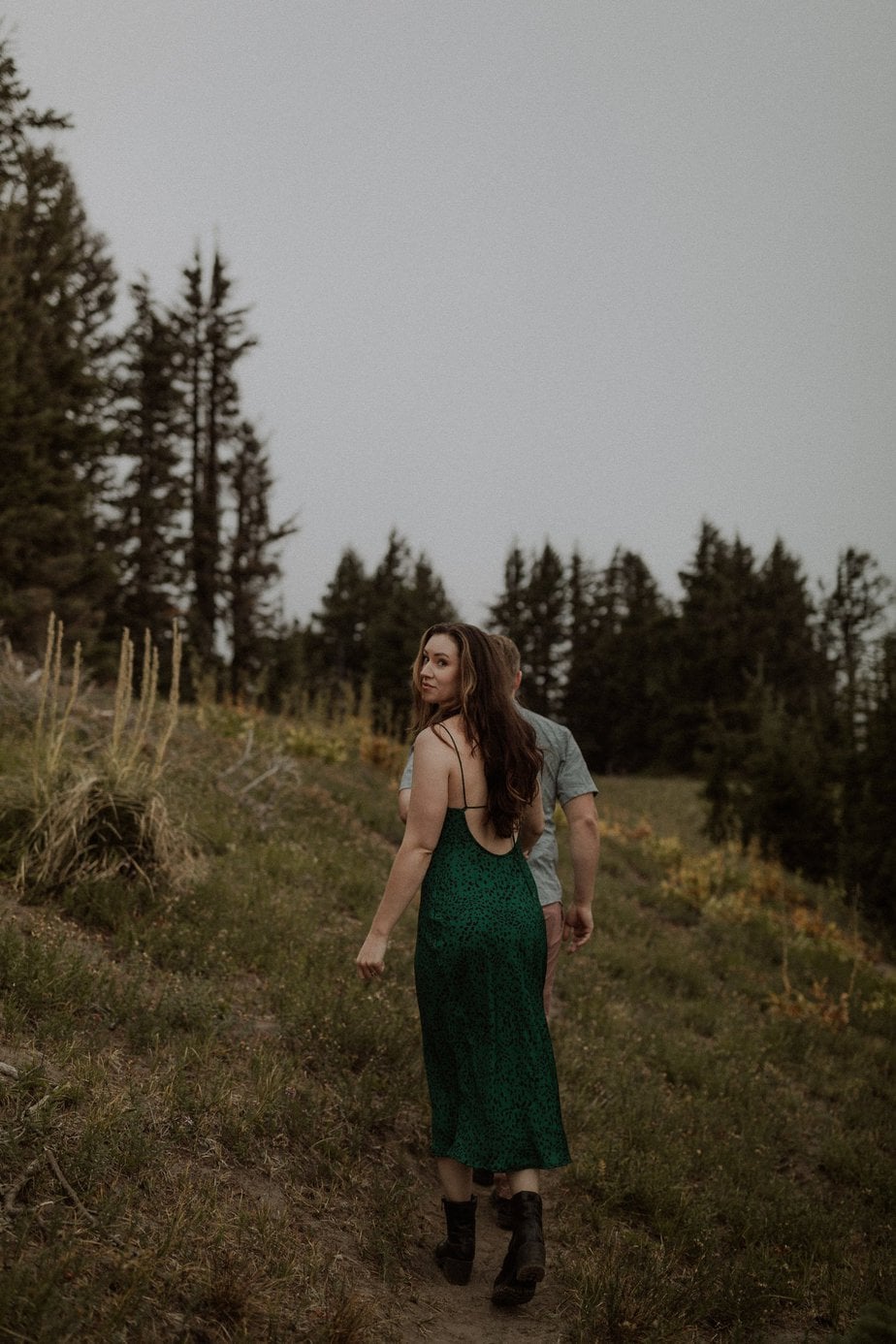 Couple walks along pathway for their Mt Hood elopement. Bride looks back over her left shoulder at photographer. She wears boots and a green dress