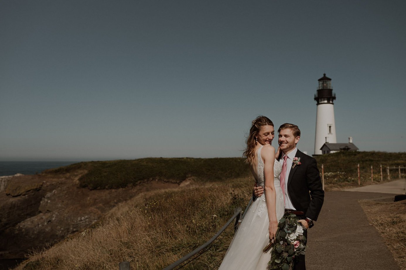 Bride and groom snuggle close at their cobble beach elopement. Yaquina Head lighthouse is in the background. Bride laughs at the groom