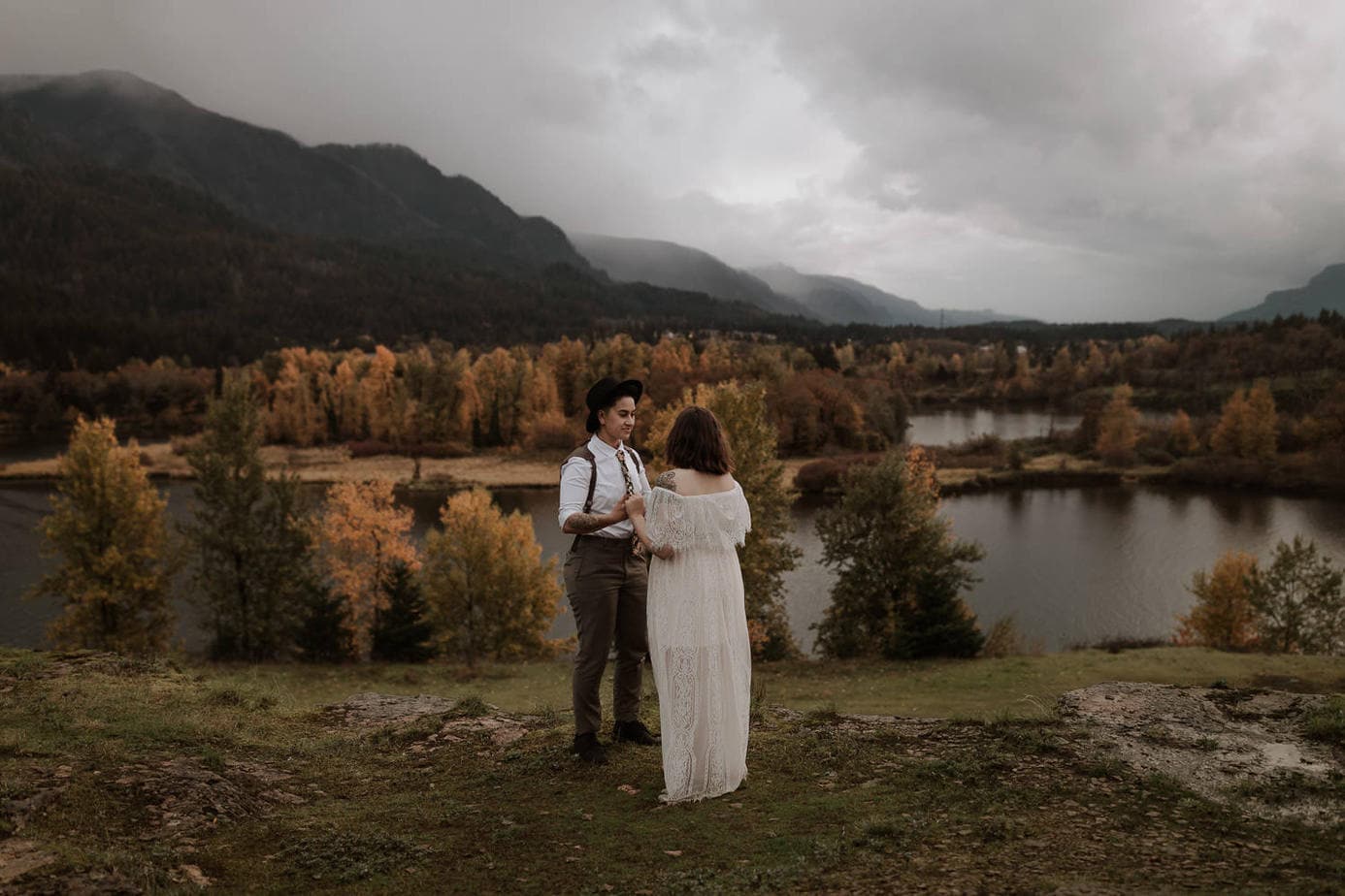 LGBTQ elopement in Oregon at the Columbia River gorge. The brides stand on the top of a cliff overlooking the river and the trees below. The trees are turning yellow from the fall season. The brides look at each other