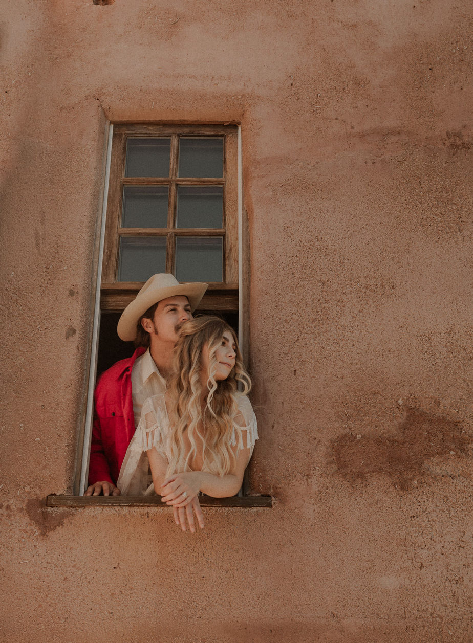 FAQ about elopements. Bride and groom hang out of window on vintage adobe in the desert. Bride's long blond hair looks like rapunzel and the groom wears a bride red jacket and cowboy hat