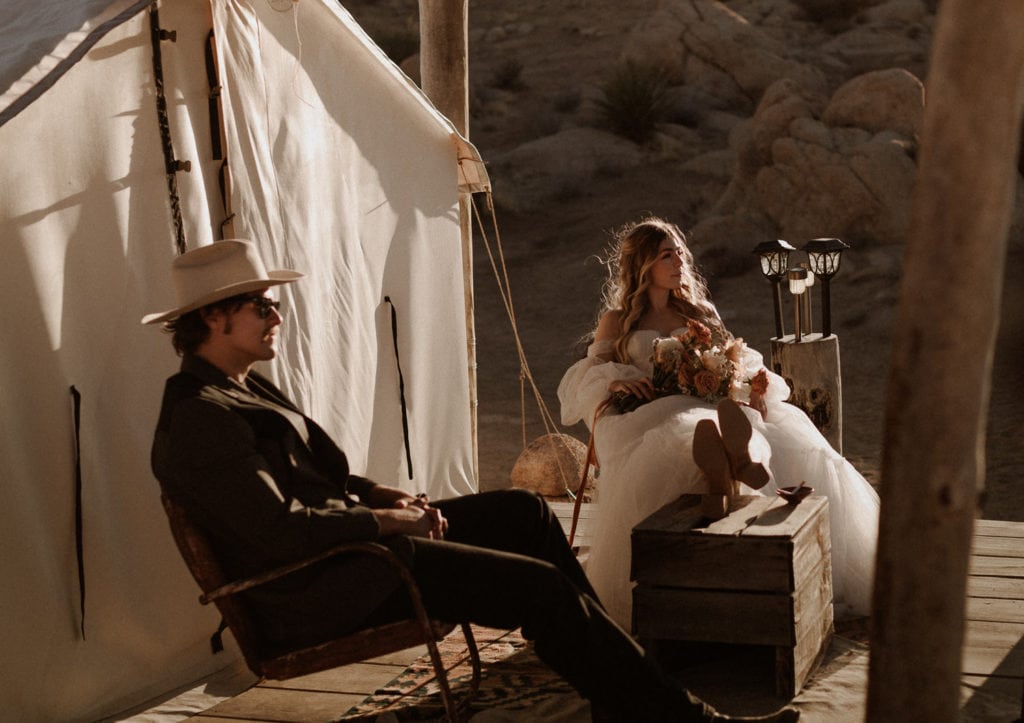 Intimate desert elopement in Joshua Tree. Bride and groom are sitting on outside their yurt while the sun goes down. Bride has her feet up on a box and the groom wears a white cowboy hat