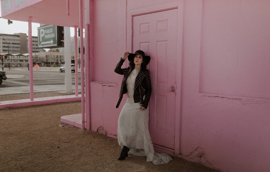 Bride leans on pink wall for bridal portraits in Las Vegas. She wears a white wedding dress, black leather jacket, and black hate