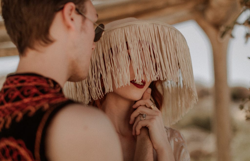Bride wears a white fringe hat and red lipstick while groom holds her face