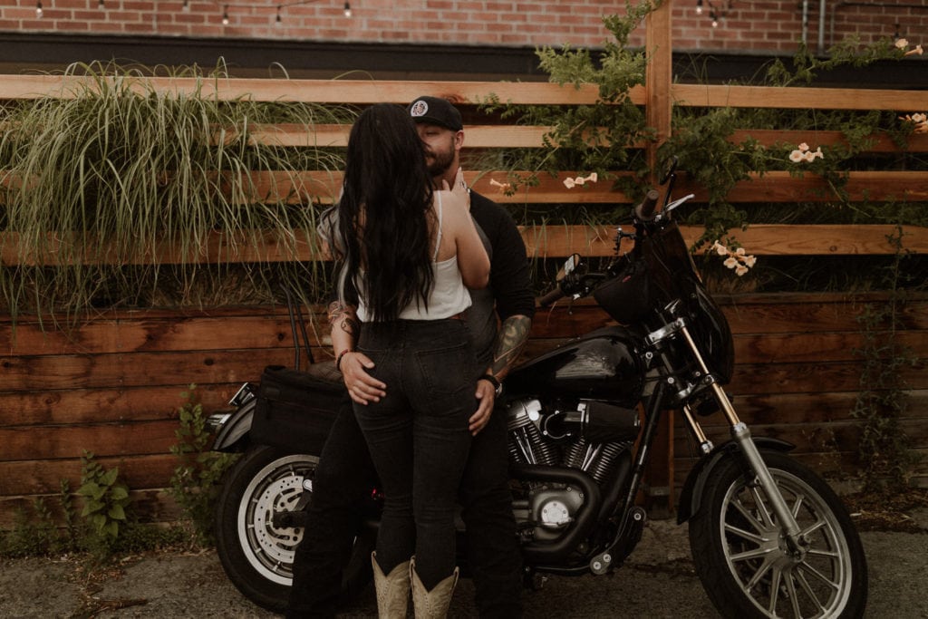 Edgy motorcycle engagement session. Guy sits on bike and his girlfriend leans on him