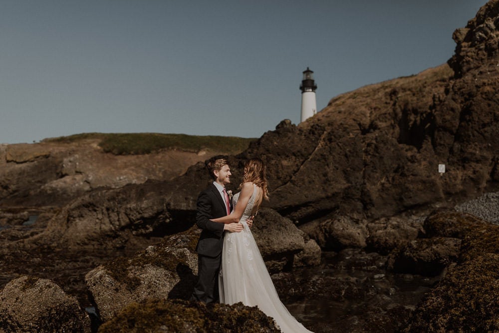 Oregon elopement photography on the coast. Wedding couple embraces on the rocks at Cobble beach with Yaquina Head lighthouse in the background