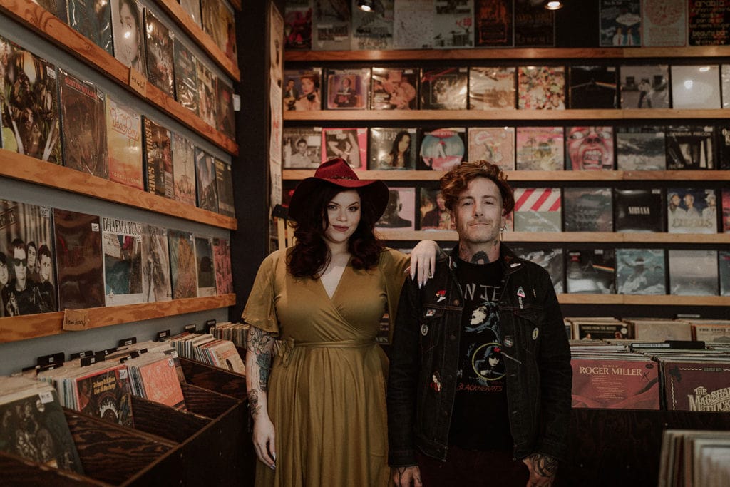 Couple stands in Vegas record shop for their engagement photos. She wears a red hat and yellow dress and he has on black leather jacket