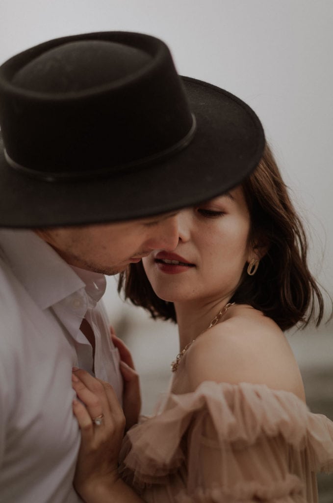 Groom wearing a black hat looks down at bride and leans in to kiss her