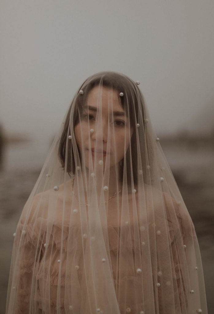 Bride looks at camera and wears a long cathedral veil that is covered in pearls