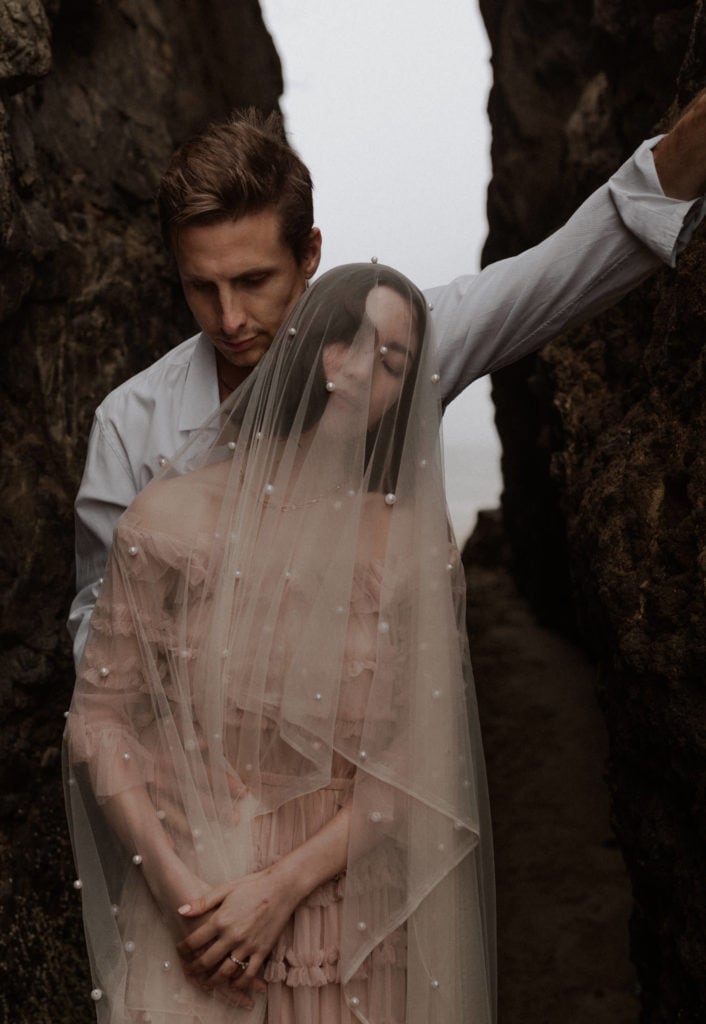 Bride and groom snuggle in Proposal rock crevice. Groom wraps his arm around her and her cathedral veil with pearls covers her face