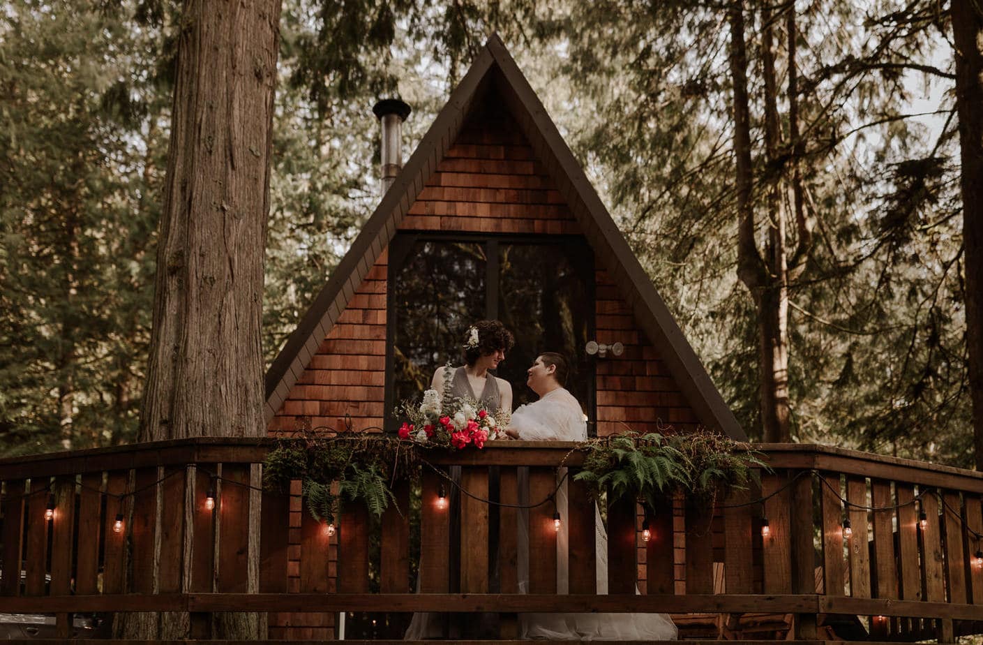 Two brides stand in front of a frame cabin in Mt Hood and look at each other. The bouquet sits on the deck ledge and the forest is behind the cabin