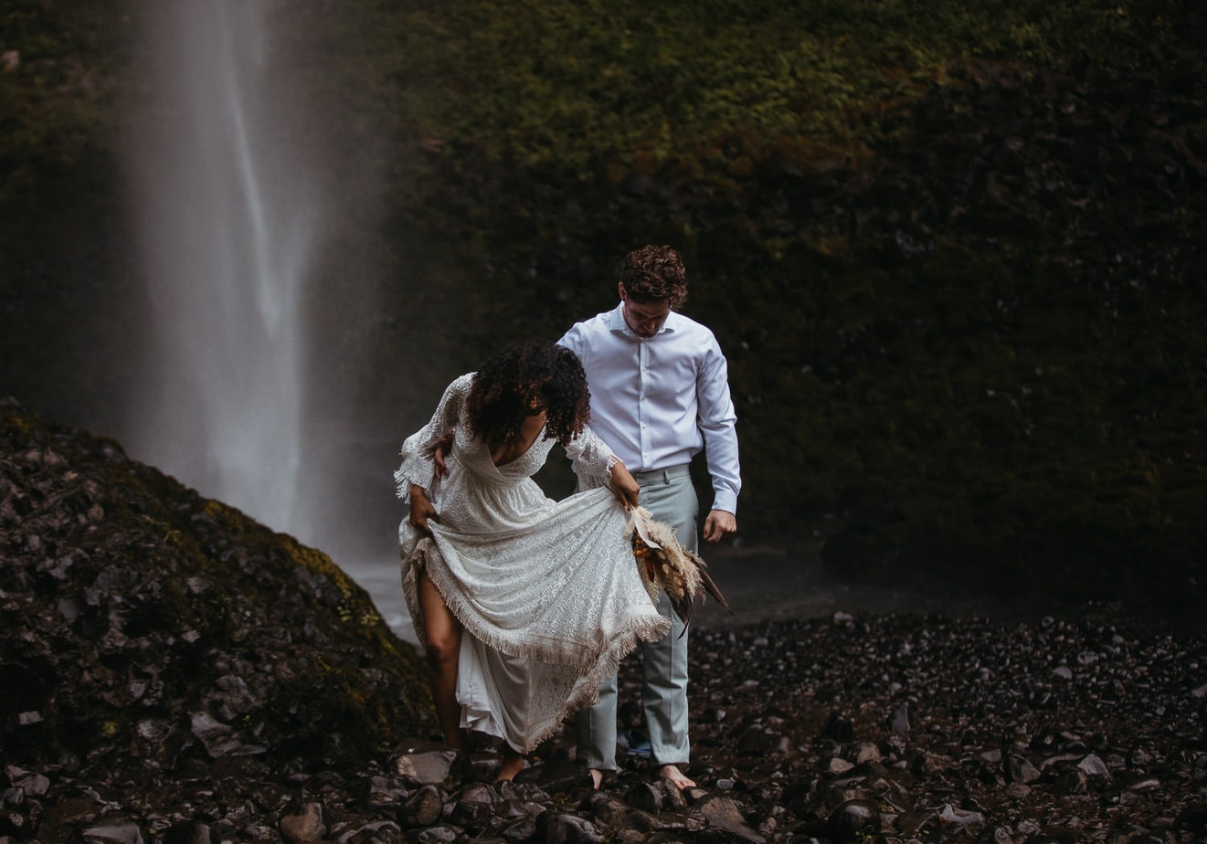 Eloping couple carefully climb over wet rocks. Bridge holds her damp dress in her hands and clutches her bouquet