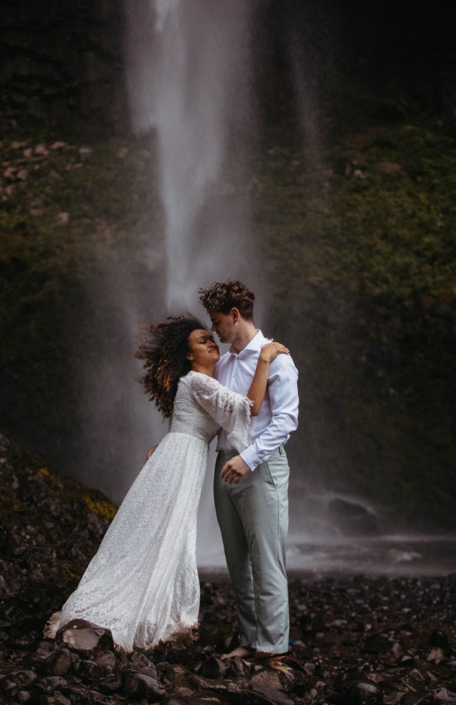 Eloping couple stand barefoot on black rock underneath Latourell Waterfall after their elopement ceremony. Bride holds onto groom's shoulder and her hair blows in the wind